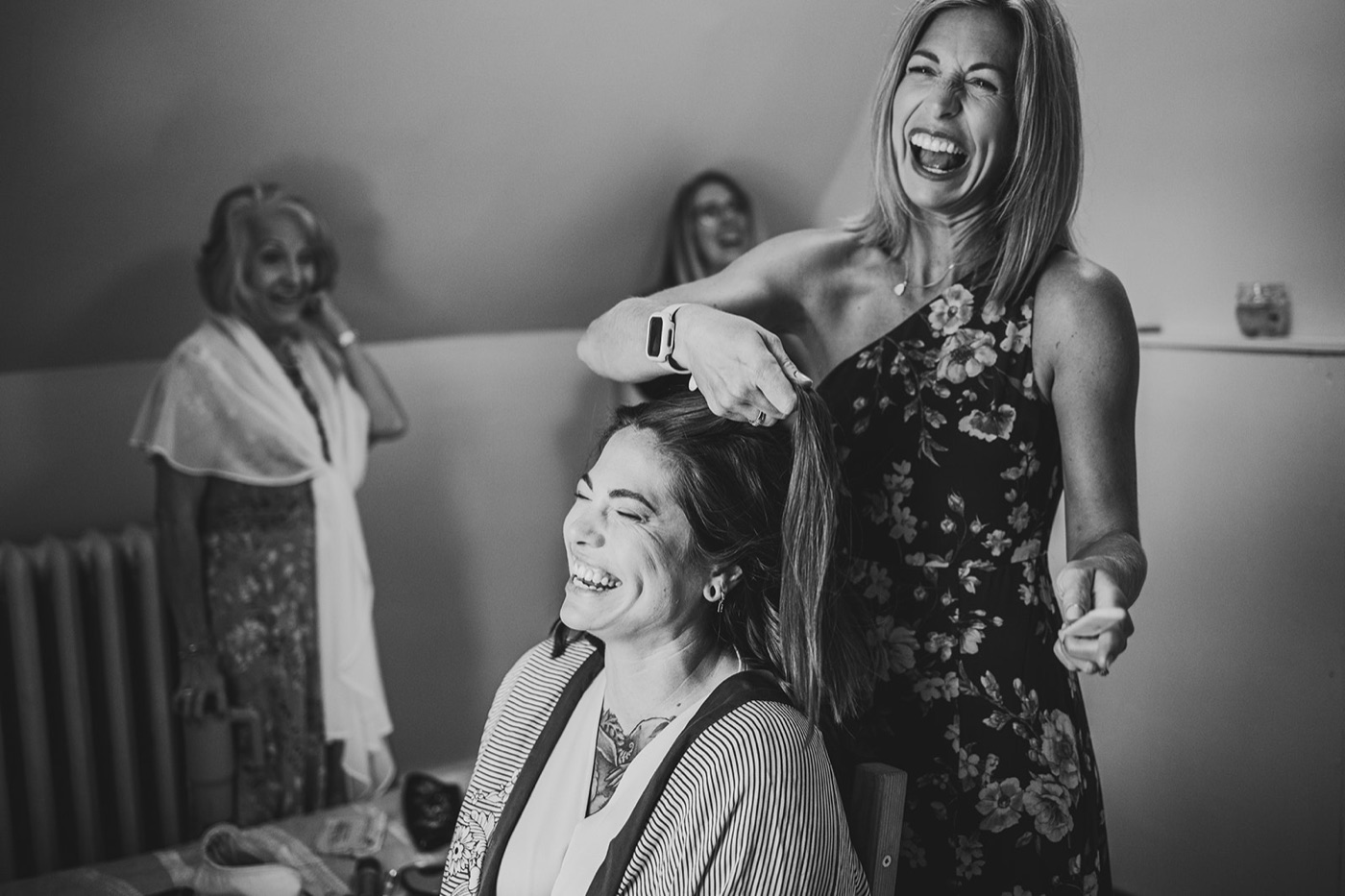 A black and white frame of a woman laughing as she styles the ponytail of a seated friend who is cracking up while getting ready