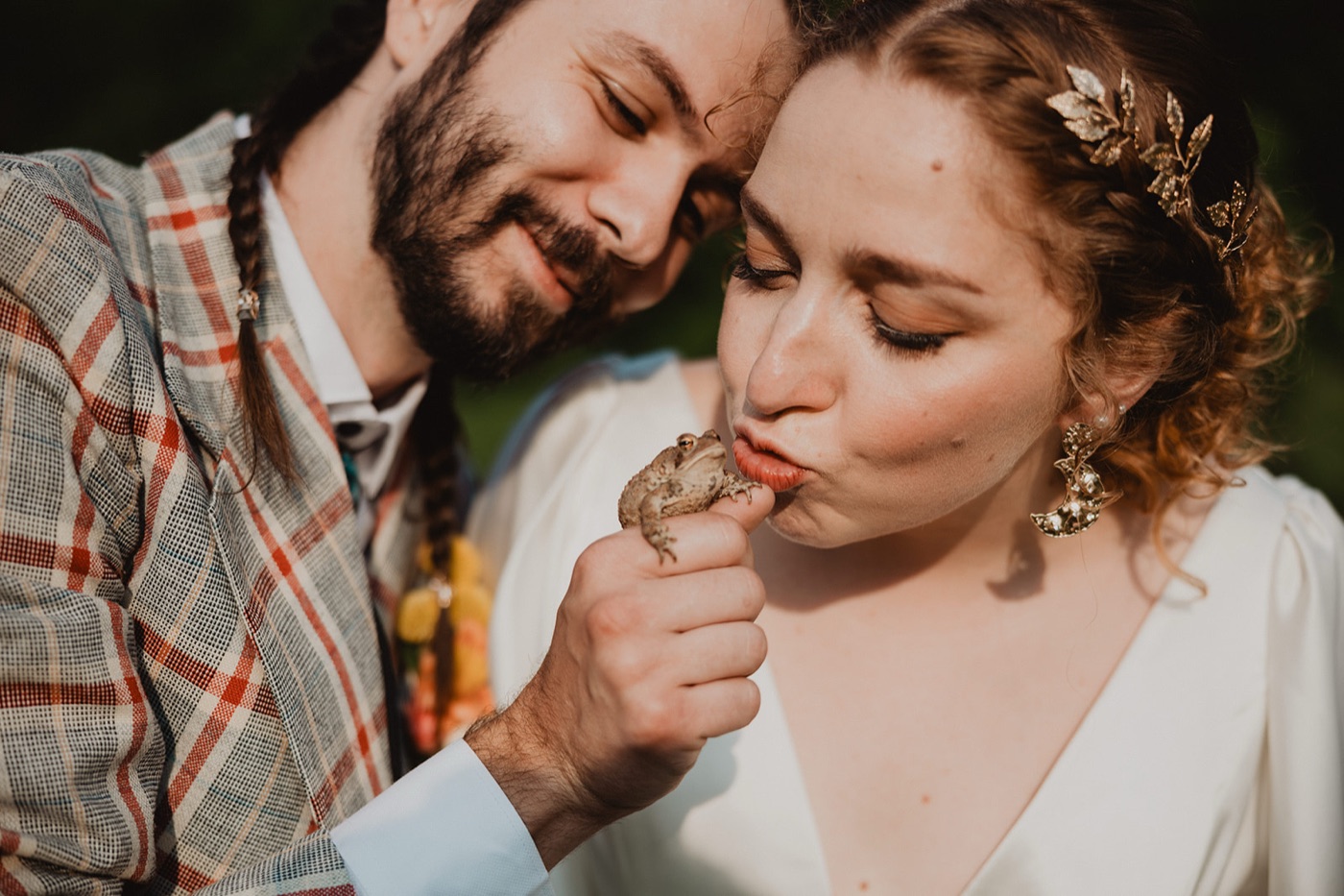 A bride puckering to kiss a toad perched on the groom's fingers as he rests his cheek against her head in warm sunlight