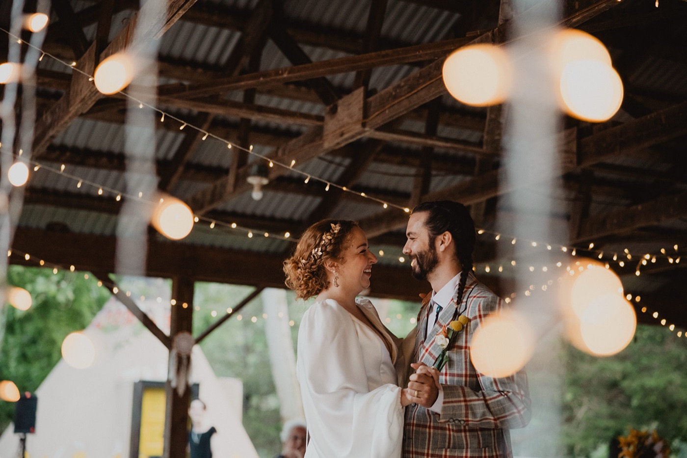 A bride and groom laughing face to face during their first dance under a timber pavilion strung with warm lights