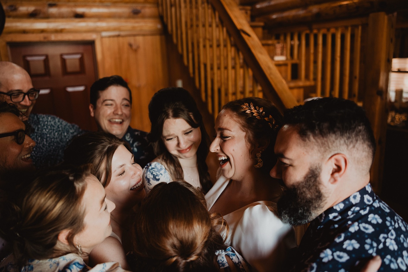 A bride laughing while wrapped in a group hug with friends and family inside a wood-paneled cabin