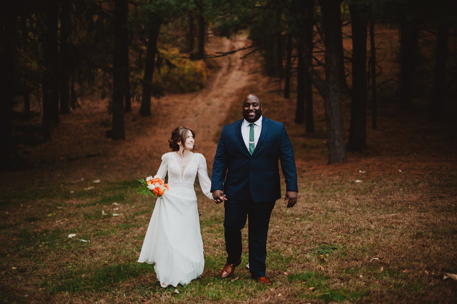 Delaney and Dante walking hand in hand out of the autumn pines, he in a navy suit and green tie, she carrying an orange bouquet