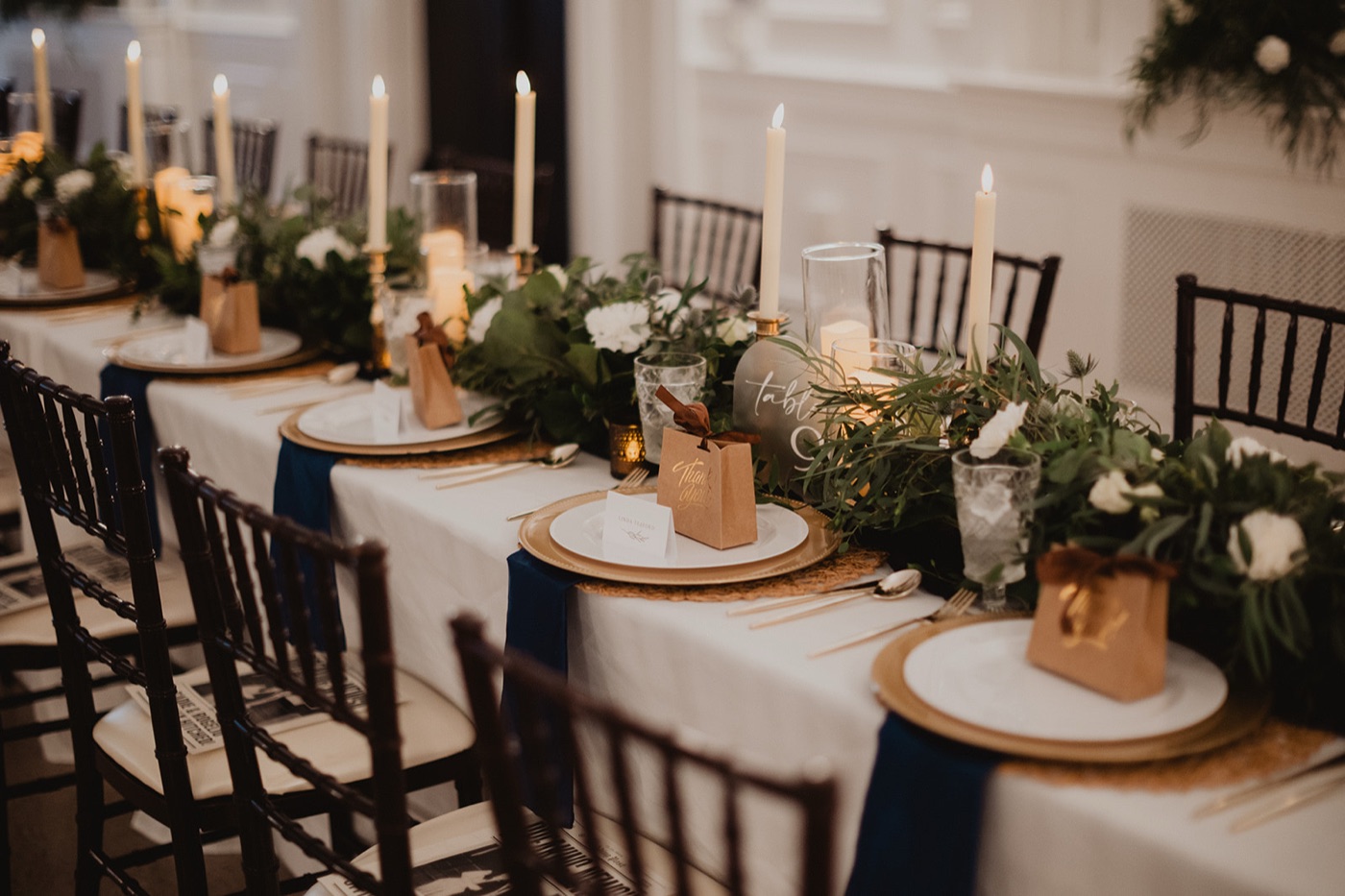 A long banquet table set with taper candles, a trailing greenery runner, gold chargers, navy napkins, and kraft thank-you favor boxes receding into candlelight