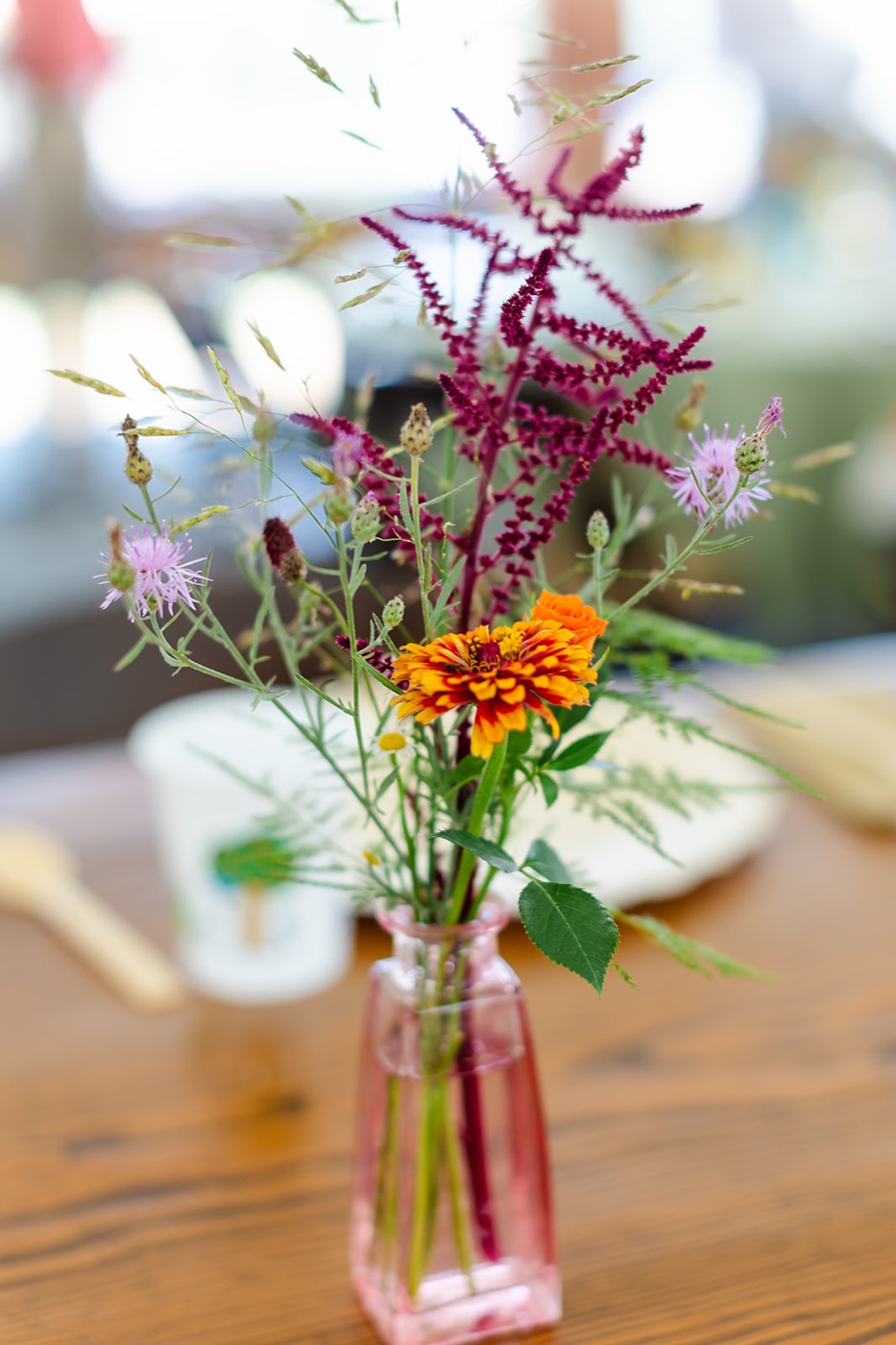 A single wildflower stem of zinnia, amaranth and grasses in a pink pressed-glass bud vase on a wood table