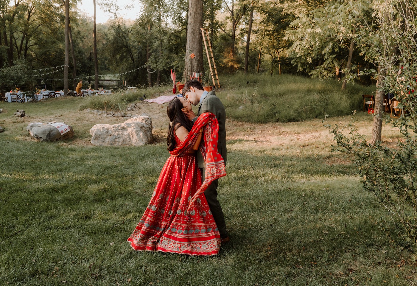 A groom dipping his bride into a kiss on a golden-lit lawn, her red block-print lehenga and dupatta sweeping out, woods and a string-lit reception behind