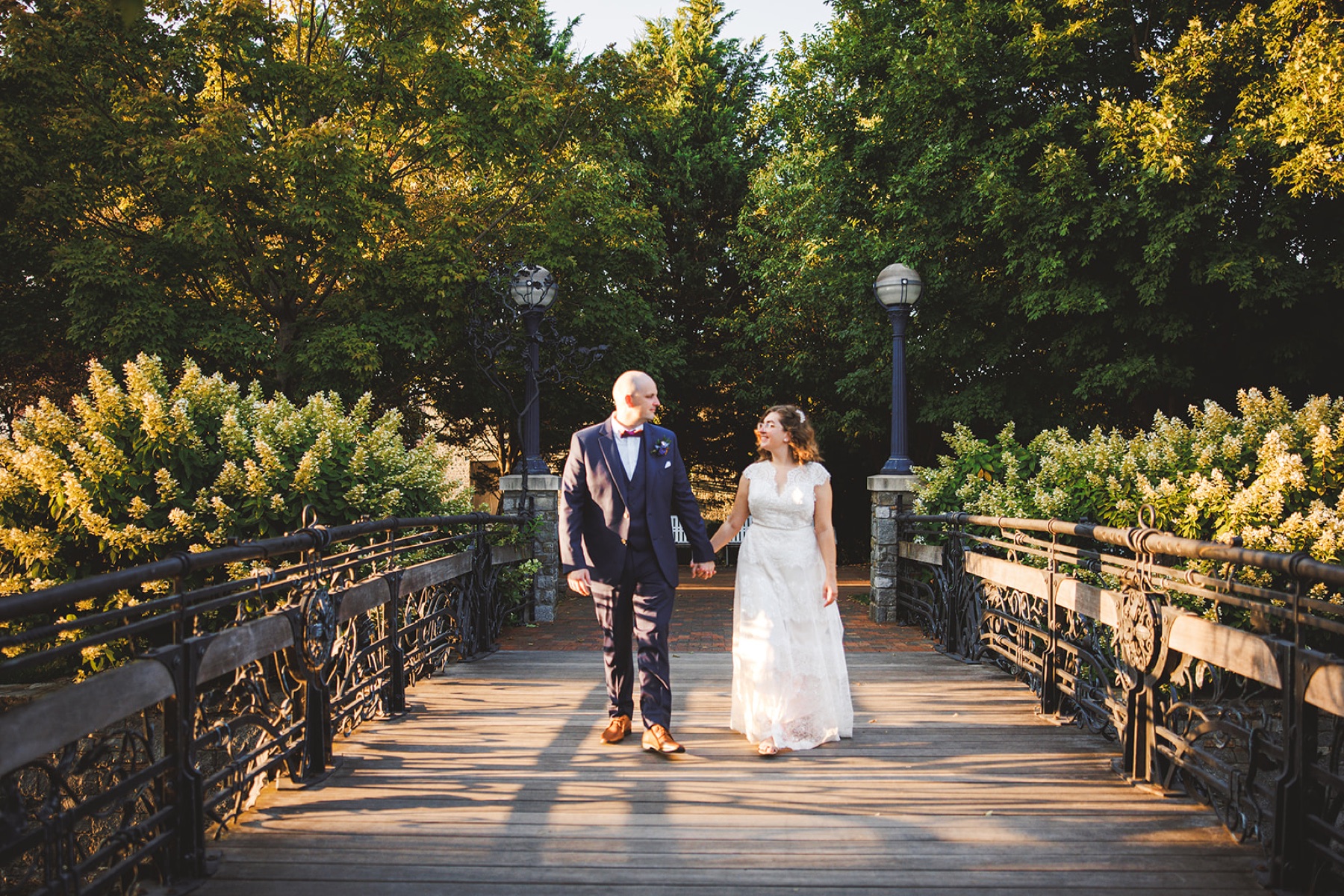 A couple walking hand in hand across an ornate iron footbridge framed by golden sunlit trees