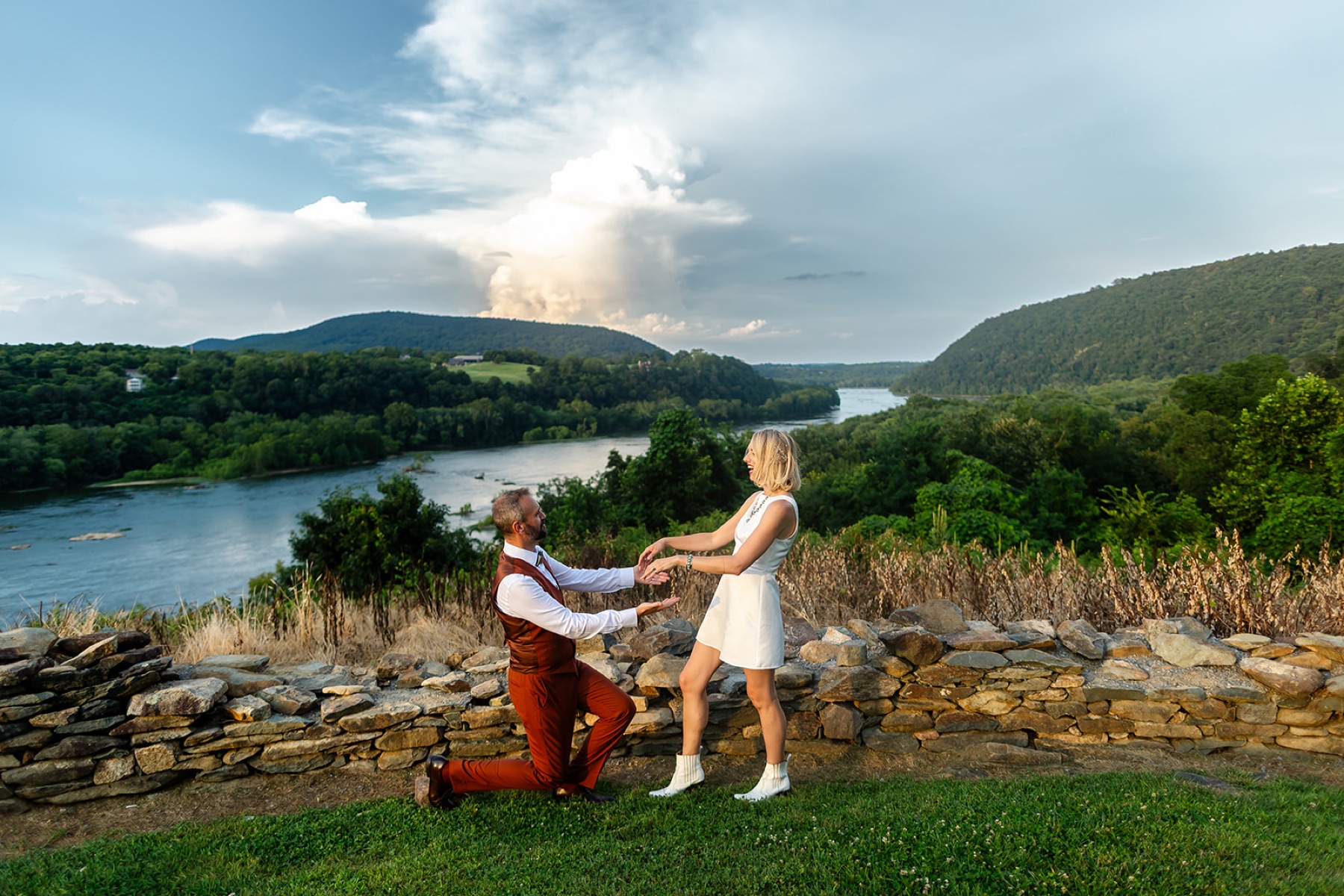 A couple on a stone overlook, the groom kneeling to take her hands above the river valley and a vast open sky